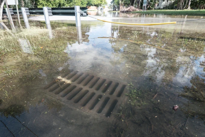 Flooded street with a visible storm drain submerged underwater. A yellow barrier stretches across the road, and vegetation lines the background, reflecting in the water.