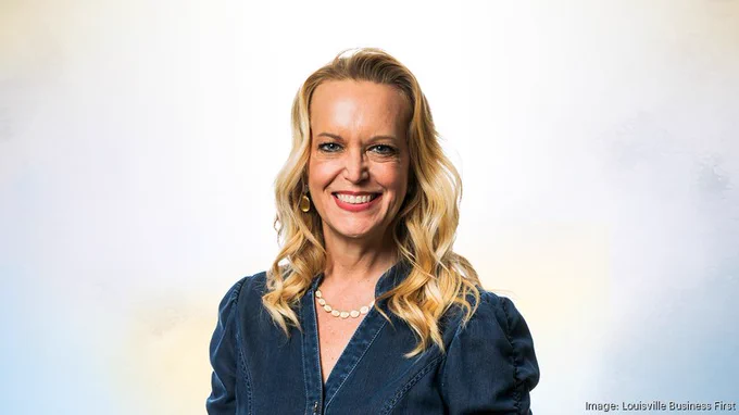 A headshot of a person with long blond hair against a white background. They are wearing a navy shirt with a v-neck collar and a white beaded necklace. They are smiling at the camera.