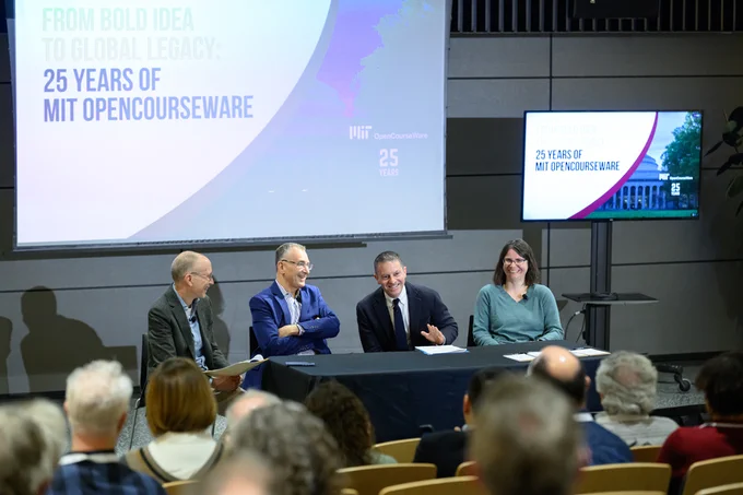 A panel of four people sits on a stage for an event celebrating 25 years of MIT OpenCourseWare. A screen displays the event's theme "From Bold Idea to Global Legacy: 25 Years of MIT OpenCourseWare." Audience members are visible in the foreground.