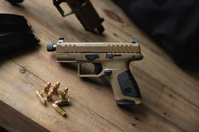 A Beretta APX pistol in a tan finish lies on a wooden surface. Nearby are several copper-colored bullets. A black object, possibly a magazine or bag, is partially visible in the corner.