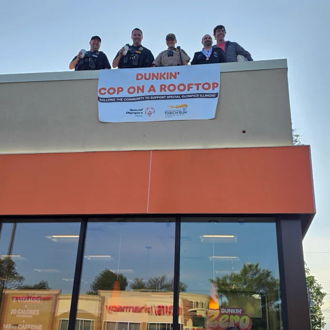 A group of individuals stand on the rooftop of a Dunkin' building, next to a banner that reads "Dunkin' Cop on a Rooftop," supporting Special Olympics Illinois.