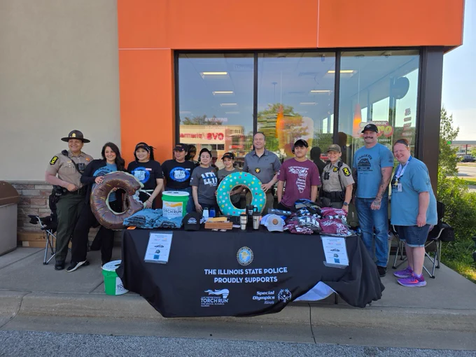 A group of people in uniform and casual clothing stand outside a building next to a table. The table is covered with a cloth that reads "The Illinois State Police Proudly Supports," featuring materials and donuts themed around "Cop on a Rooftop" and "Special Olympics Illinois." The setting appears to be a charity event.
