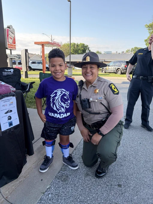 A person in uniform kneels next to a smiling child wearing a purple shirt. They are outdoors near a table with materials displaying the Dunkin' logo. Other people in uniform stand in the background.
