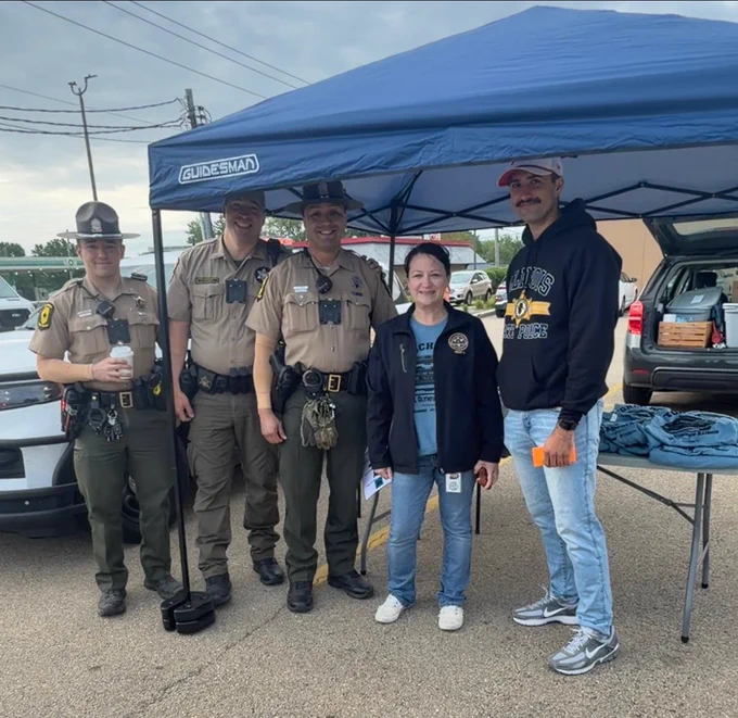 A group of five people stands under a blue Guidesman canopy next to parked vehicles. Three individuals wear law enforcement uniforms. There is a table with items on display beside them.