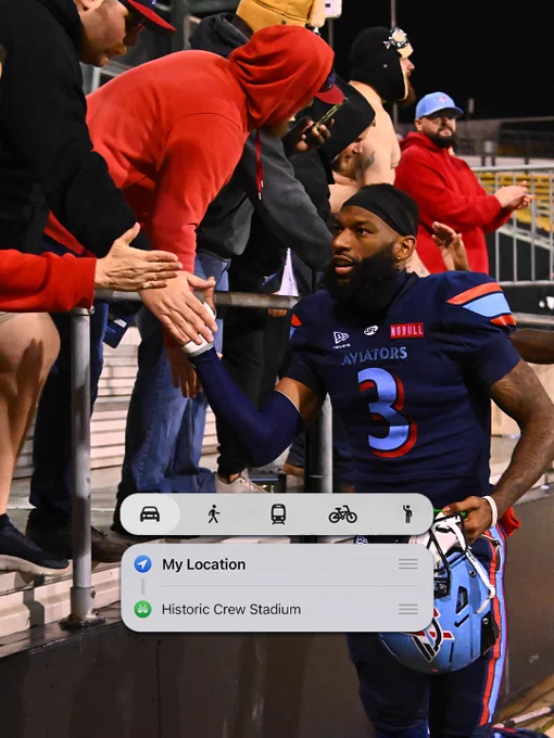 A football player in a blue uniform shakes hands with fans standing in stadium bleachers. The mood is energetic and appreciative.