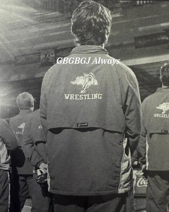 A person stands with their back to the camera, wearing a jacket labeled "Wrestling" with a jackrabbit logo. The text "GBGBGJ Always" is written above. Others in similar attire stand nearby in an indoor setting.