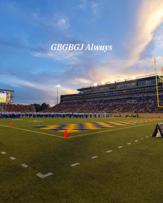 A large football stadium is filled with spectators, with a football field in the foreground featuring markings and goalposts. A marching band, dressed in blue and white, is performing on the field. The sky is dramatic with a mix of clouds and sunset colors. A scoreboard and stadium lights are visible. "GBGBGJ Always" is written in the sky.