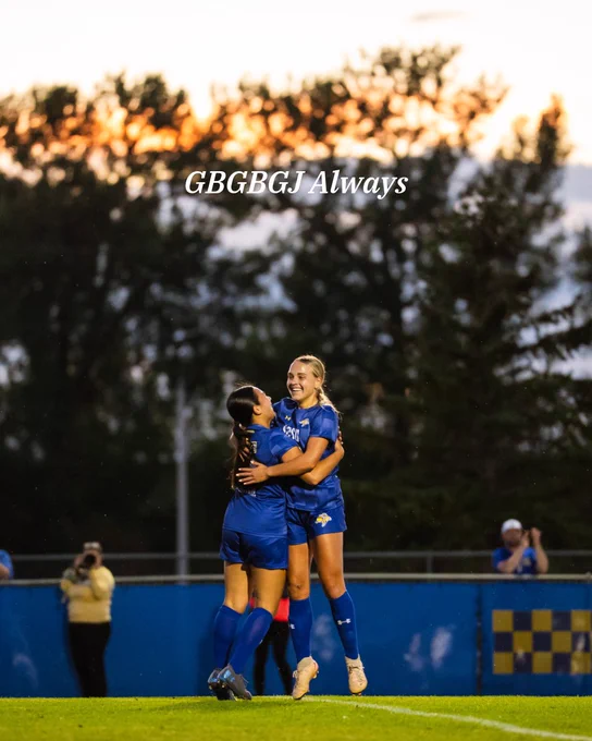 Two soccer players in blue uniforms celebrate on the field, embracing each other. The sky is orange and dark blue, indicating sunset. In the background, a few spectators watch, and trees are silhouetted against the sky. The phrase "GBGBGJ Always" is written across the top of the image.