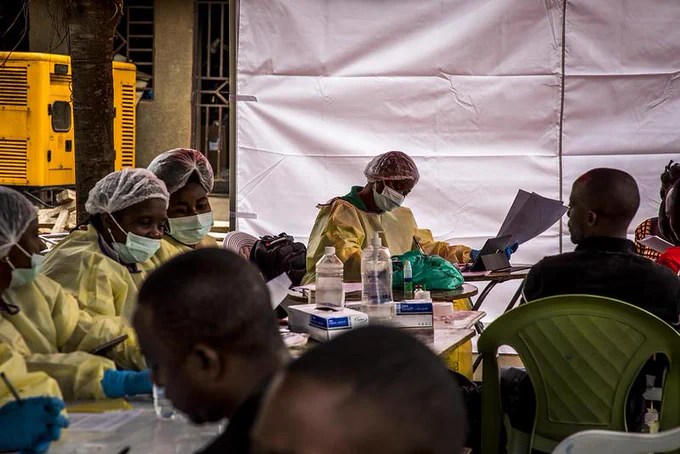 Ebola doctors in protective gear reviewing documents at a makeshift clinic with seated patients nearby.