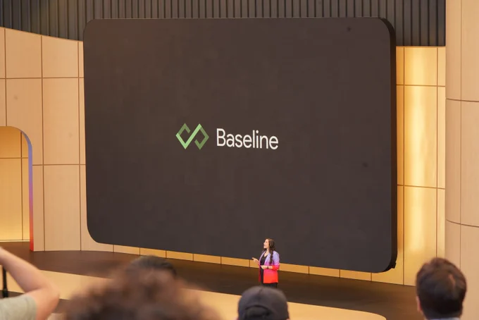 A wide stage shot shows a presenter standing at the center of the stage in front of a giant dark screen displaying a green infinity-style geometric logo next to the word "Baseline." The presenter is a woman with long dark hair wearing a vibrant purple-to-pink gradient blazer over black clothing.