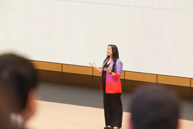 A medium shot from the audience's perspective shows a female presenter speaking with her hands outstretched. She has long, dark wavy hair and is wearing a striking purple, pink, and bright red gradient blazer over a black top and wide-leg trousers.