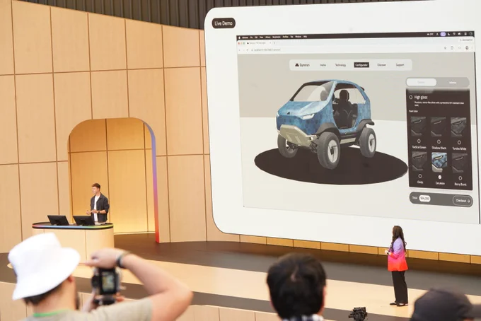 A wide stage shot capturing a live demo on a large screen showing a web-based 3D vehicle configurator interface featuring a small blue off-road electric car. A male presenter stands at a wooden podium on the left side of the stage, while the female presenter in the gradient blazer watches from the right side. In the foreground, blurred audience members, including one wearing a white bucket hat, look on.
