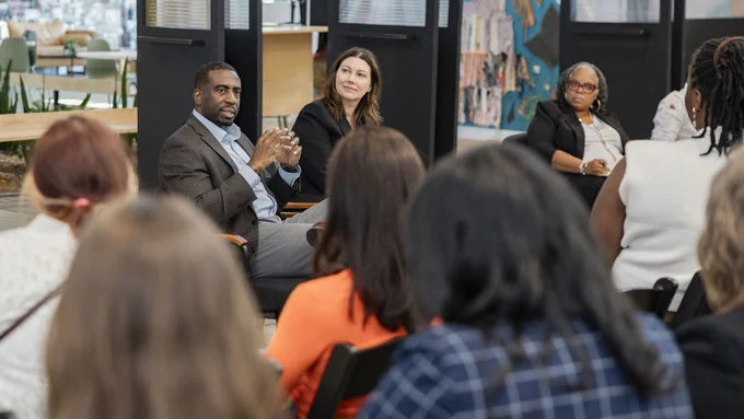 A diverse group of people seated in a meeting featuring a man speaking to an audience, viewed over the shoulders of attendees in a modern office setting.