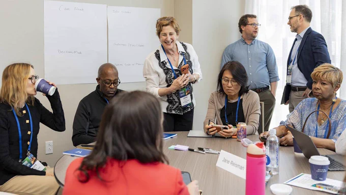 A diverse group of professionals attends a meeting around a table with a smiling woman standing near a whiteboard listing notes.