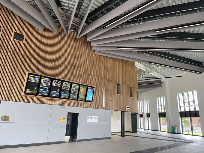 New open concourse with radial steel ceiling beams and train display boards on a wood strip clad wall.