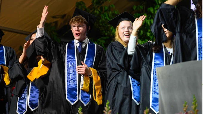 A group of graduates in black caps and gowns stands together, some raising their arms in celebration. They wear blue stoles with "MSFS" embroidered on them. The background features green plants and a tent.