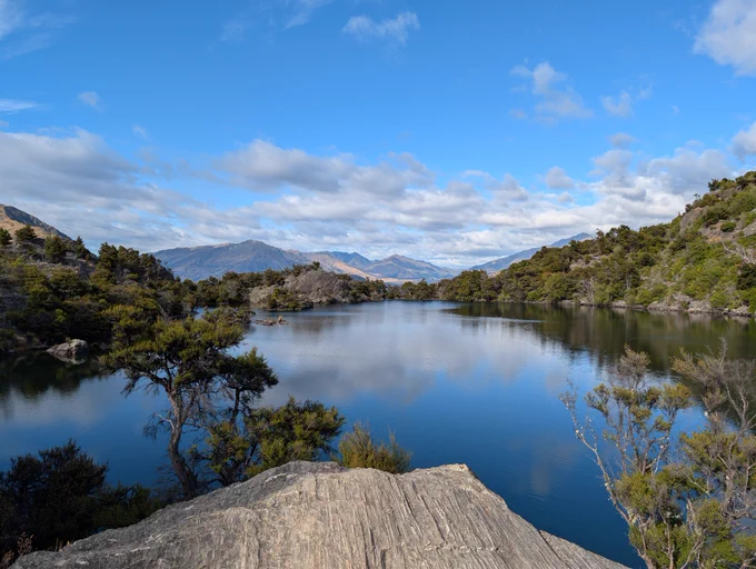 A photo of a lake reflecting a partially cloudy sky and surrounded by mountains.