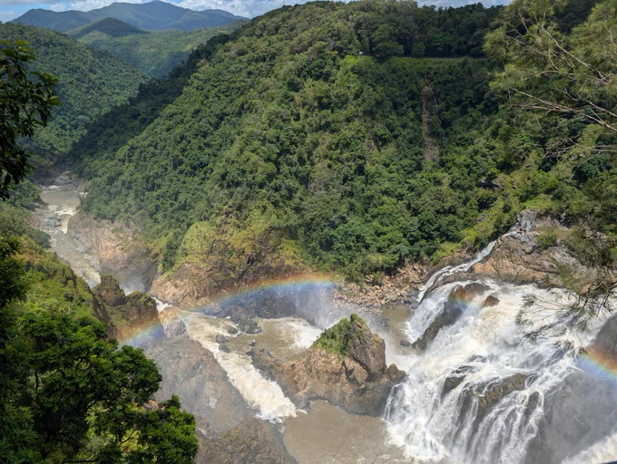 A photo overlooking a waterfall cutting through rainforest covered mountains and into a river running away from the photographer. A rainbow crosses over the waterfall.