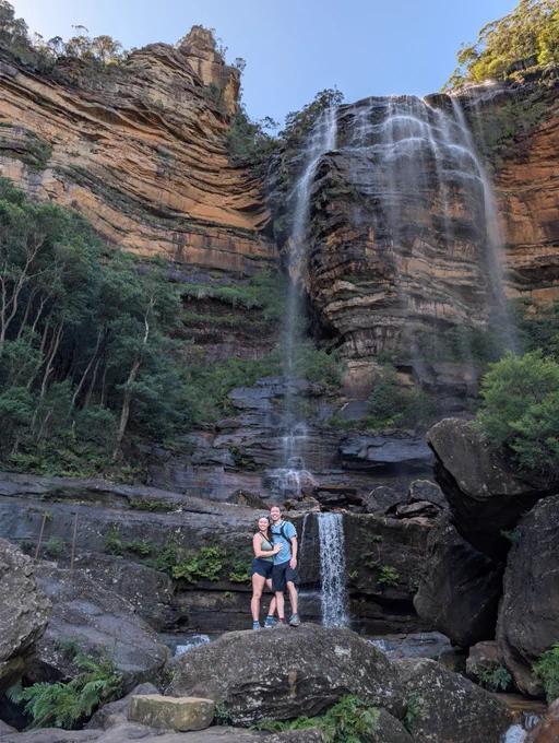 A photo of Tyler and Kira standing in front of a waterfall. The photo is distant and angled to show the height of the rocky cliff the water flows over.