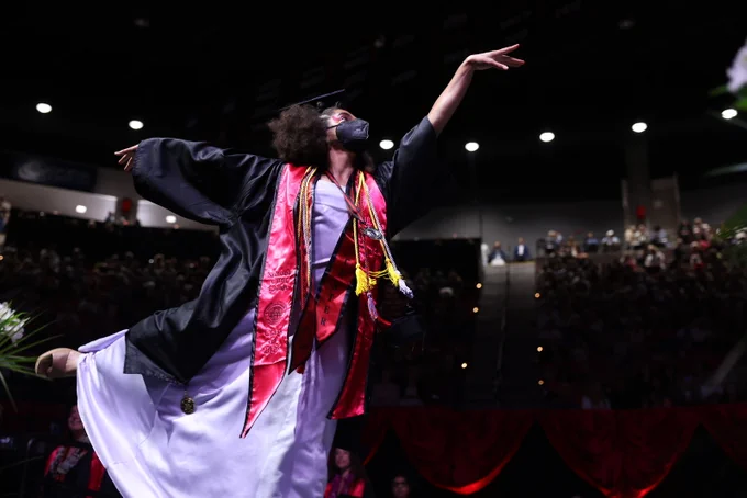 A person in a graduation gown and cap performs a graceful dance move on stage during a ceremony. They are wearing a decorative stole and cords, with a large audience in the background.