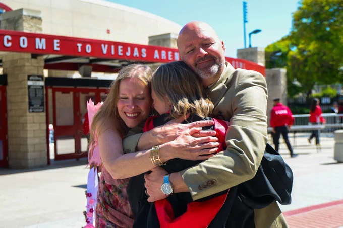Three people are embracing happily outside Viejas Arena, celebrating a graduation. One is in regalia, holding a diploma. The scene is joyful with a sunny backdrop.