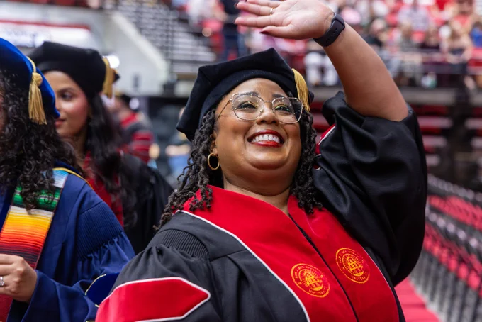 A person in academic regalia smiles and waves during a graduation ceremony. The robes feature red and black with the San Diego State University logo. A crowded auditorium is in the background.