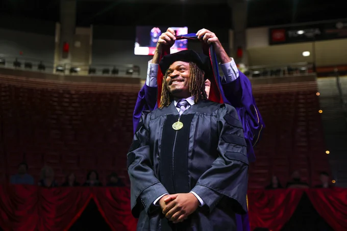 A graduate in a cap and gown smiles while someone adjusts their cap during a ceremony. There's a gold medallion around the graduate's neck.