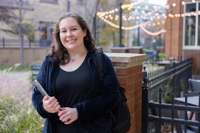 A CSU student standing outside the administrative building smiling with a textbook and backpack. 