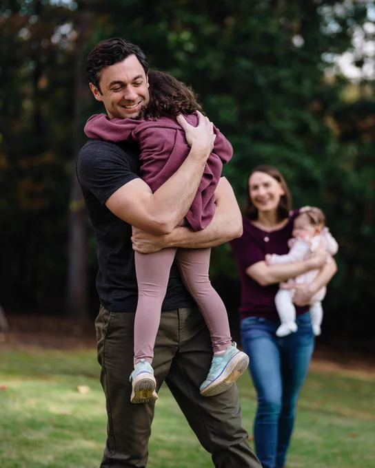 A photo of Senator Jon Ossoff with his family outdoors. He is holding his daughter in his arms.