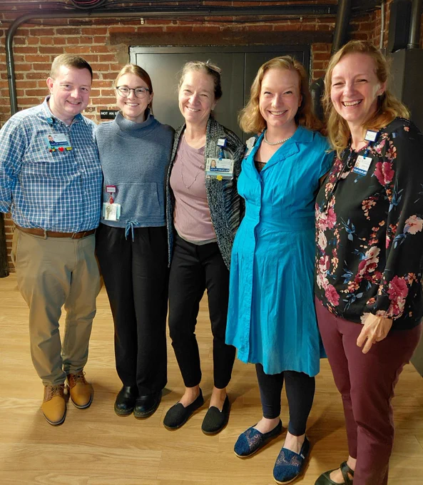Five smiling individuals stand shoulder-to-shoulder against a brick wall. From left to right: SCOTT, CHAPLAIN ASSOCIATE; a person wearing glasses; PAM, STAFF CHAPLAIN; a person in a blue dress; and KRISTINA, STAFF CHAPLAIN.