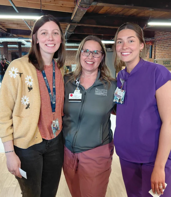 Three smiling healthcare workers stand shoulder-to-shoulder. The woman in the center wears glasses and a jacket labeled \"PALLIATIVE MEDICINE CONSULTANTS.\" Lanyards with \"Penn Medicine Princeton Health\" and ID badges are visible. \"30 DOZEN EGGS\" is partially visible.