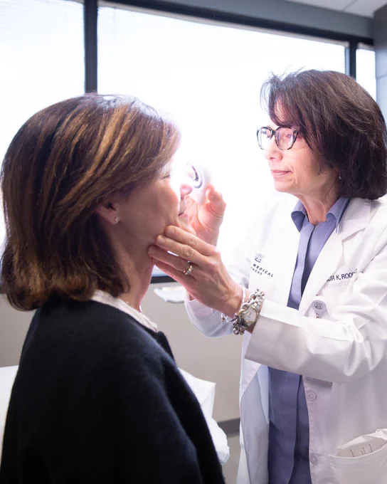 A healthcare professional in a white coat examines a person's face using a small circular instrument in an office setting with natural light coming through the windows.