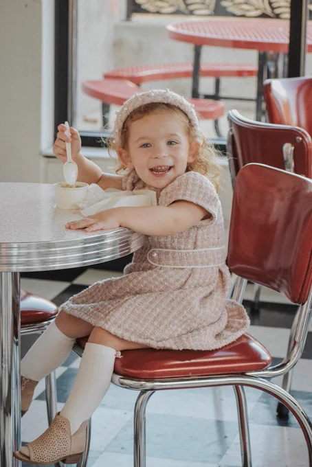 A child with curly hair sits on a red diner chair, smiling while holding a spoon over a cup of ice cream at a table. In the background, there are more red chairs and a window with decorative patterns.