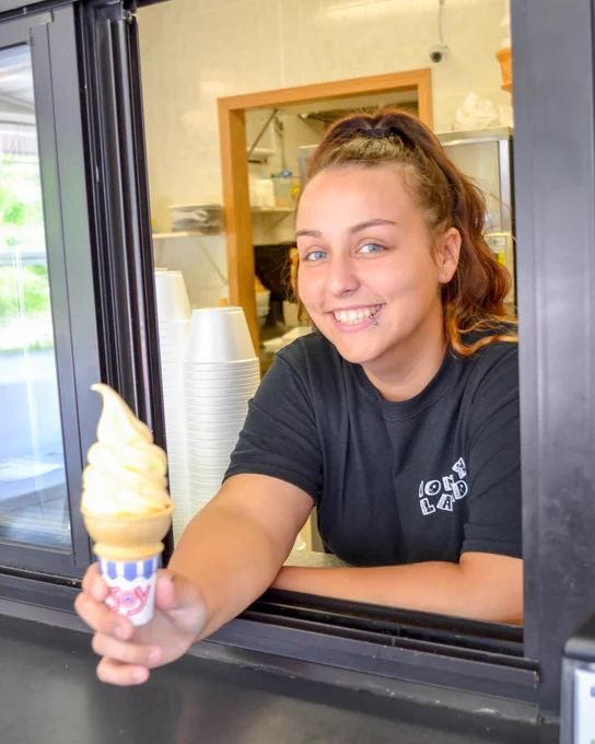 A person serving a soft-serve ice cream cone through a takeout window at Coney Land. The ice cream is swirled high above the cone. Stacks of cups are visible in the background.
