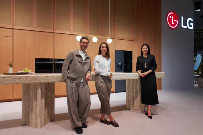LG brand showroom featuring three people standing in a modern kitchen display area with built-in LG appliances, wood-paneled cabinetry, a stone island counter, pendant lights, and an illuminated LG logo on the wall.