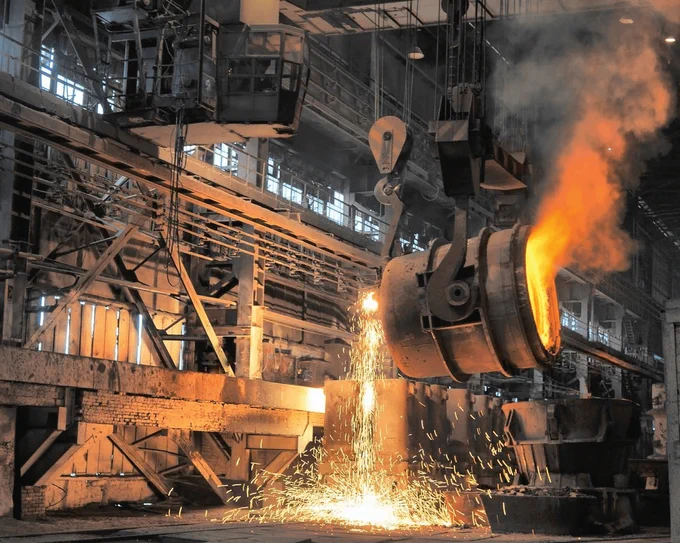 Molten aluminium pours from a large crucible in an industrial setting, with sparks flying and intense heat visible.