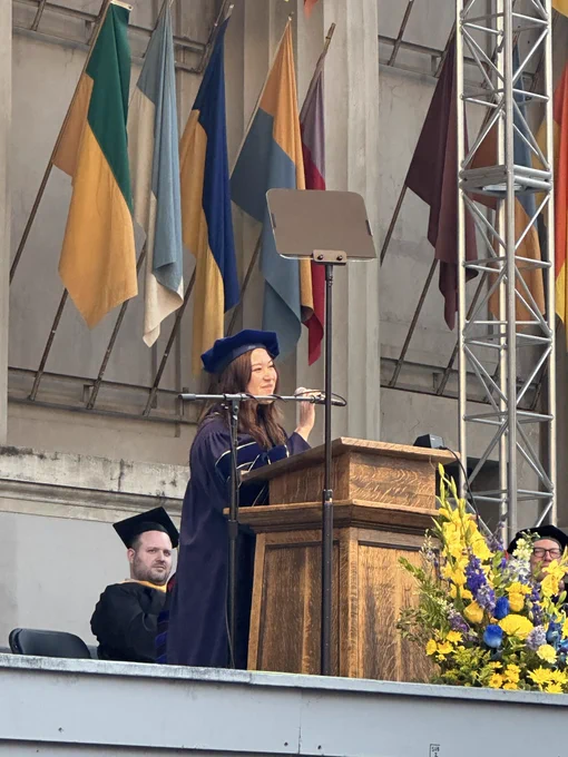Woman in blue regalia holds microphone at lecturn with blue and yellow flowers in the front and flags in the back