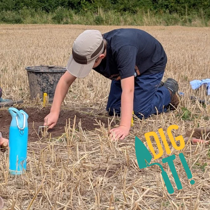 A young person digs on an archaeological excavation. They are kneeling in a ploughed field and scraping soil with a trowel.