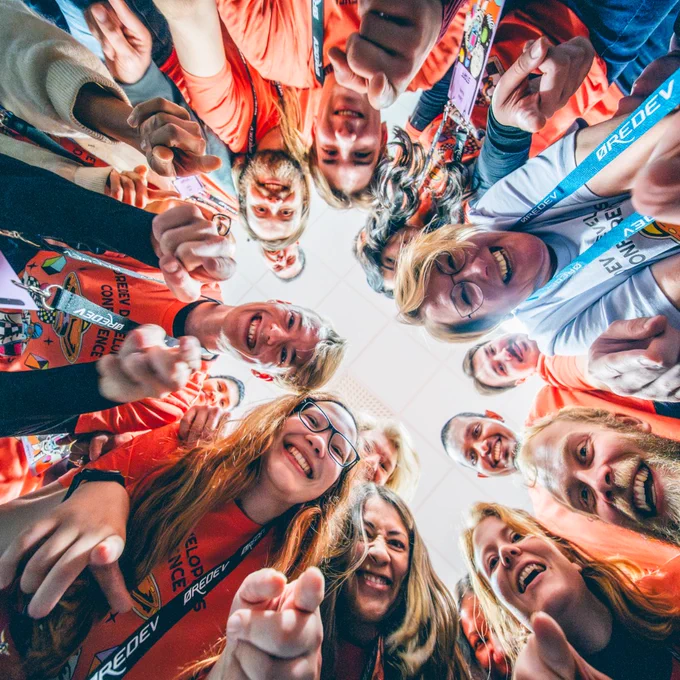 Group of smiling volunteers in colorful volunteer t-shirts forming a circle and pointing toward the camera in a joyful team moment.