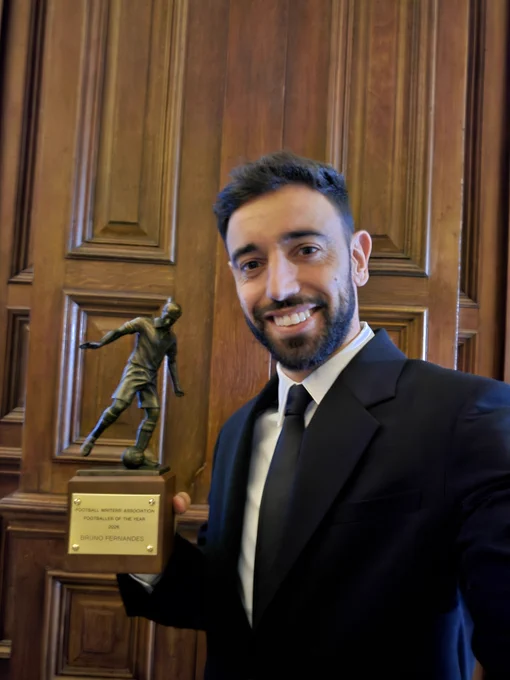 Bruno Fernandes wearing a dark suit, holding the FWA Men's Footballer of the Year trophy with a wooden paneled background and smiling as he takes a selfie.