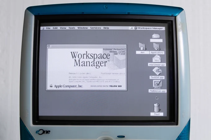 A medium close-up of a blue and white eMachines eOne all-in-one computer. The CRT monitor shows a grayscale Rhapsody desktop with a menu bar on top and several icons on the right. An open window says Workspace Manager and at the bottom it says Apple Computer Inc. Developed with Yellow Box.