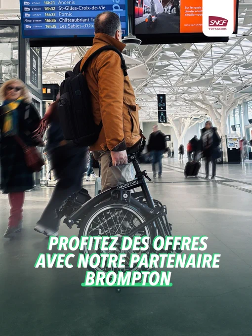 Un voyageur tenant un vélo pliant Brompton dans une gare animée avec un panneau d'affichage des trains en arrière-plan.