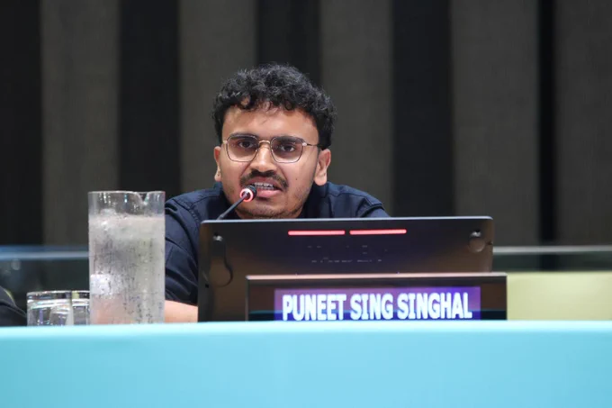 Puneet Singh Singhal speaks into a microphone while seated at a conference table with a nameplate displaying his name in front of him.