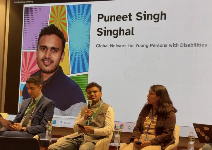 I am speaking on a panel at a conference, holding a microphone while seated between two fellow panelists. Behind us, a large screen displays my portrait, name, and affiliation with the Global Network for Young Persons with Disabilities.