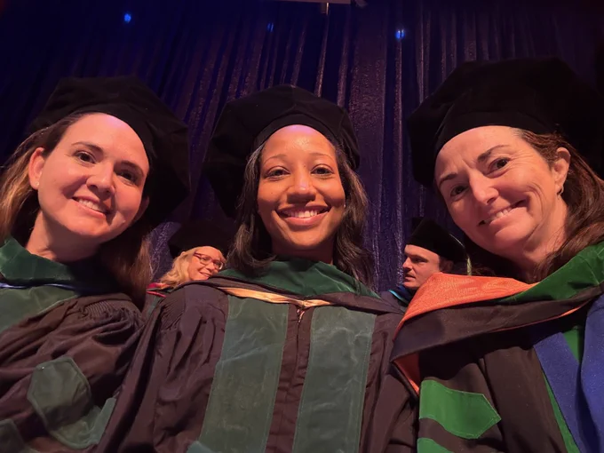 Selfie: three smiling faculty, all women, in formal black academic gowns with green trim; they are also wearing black doctoral tams on their heads