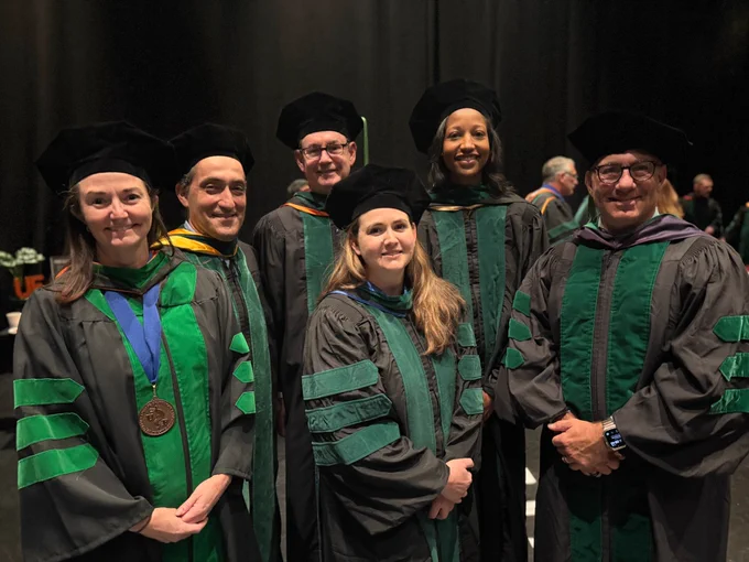  A group of six University of Florida faculty in formal black academic gowns with green trim; they are also wearing black doctoral tams on their heads.