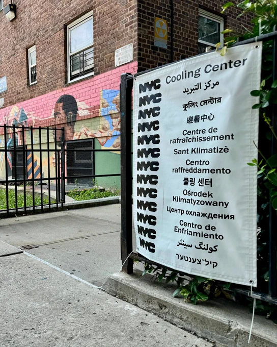 Photo of a sign for an N Y C Cooling Center in multiple languages attached to a black fence outside a brick building with a colorful mural.