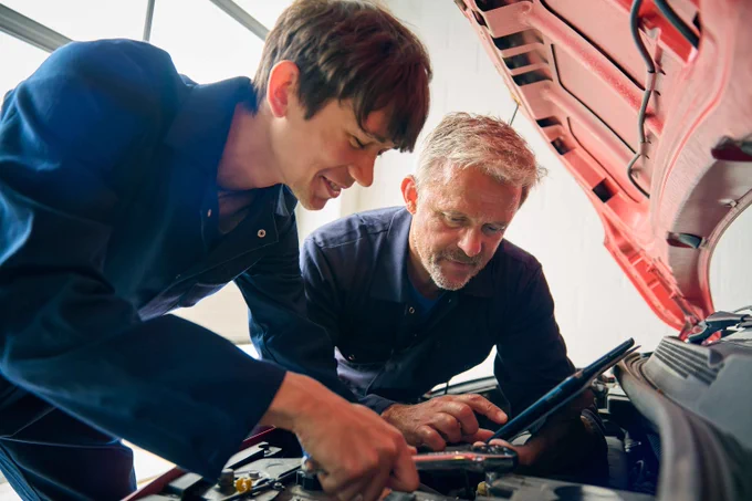 Two mechanics in blue coveralls inspecting a car engine using a tablet and tools under the open hood of a red vehicle