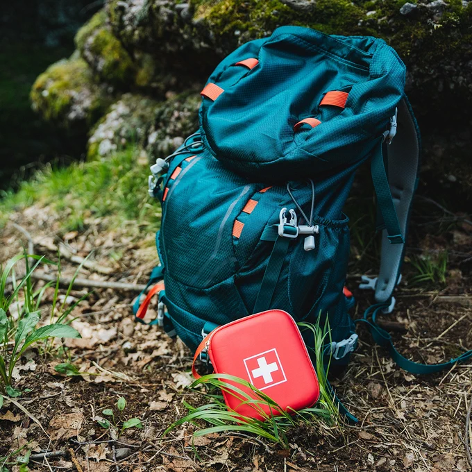 A teal hiking backpack is resting on a forest floor. In front of it, there is a red first aid kit with a white cross. Moss-covered rocks and grass surround the area.