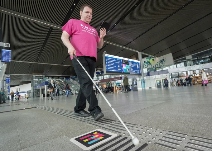Person with a white cane wearing a RNIB \"See differently\" t-shirt, using NaviLens on a tablet at Belfast Grand Central station. 